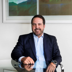 Fotografía a color de medio cuerpo del Dr. José Gregorio Rojas que se encuentra sentado en una mesa frente a una pared blanca con cuadros